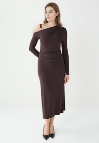 Braunes, schulterfreies Langarmkleid mit körperbetonter Silhouette, Raffung in der Taille und glatter, eleganter Textur. Schwarze High Heels.