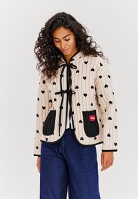 Prešívaná bunda s kapucňou z krémovej látky, so vzorom čiernych srdiečok a kontrastnými čiernymi vreckami. Obsahuje šnúrkovú zarážku a ribované manžety.