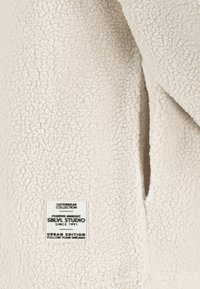 Off-white fleece stof met een zachte textuur. Bevat een label met "OUTERWEAR COLLECTION" en "SBLVLL STUDIO" branding.