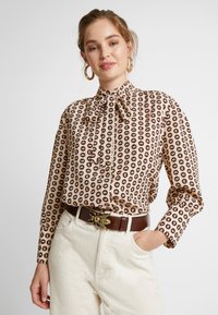 Beige Bluse mit braunem Tränenmuster, hohem Kragen und Schleife. Lange Ärmel mit Knopfmanschetten, kombiniert mit einem braunen Gürtel und cremefarbener Cordhose.