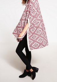 Strikket poncho i bordeaux og hvid med diamantmønstre. Kanter er kantet med hvidt. Bæres over sorte leggings og ankelstøvler.