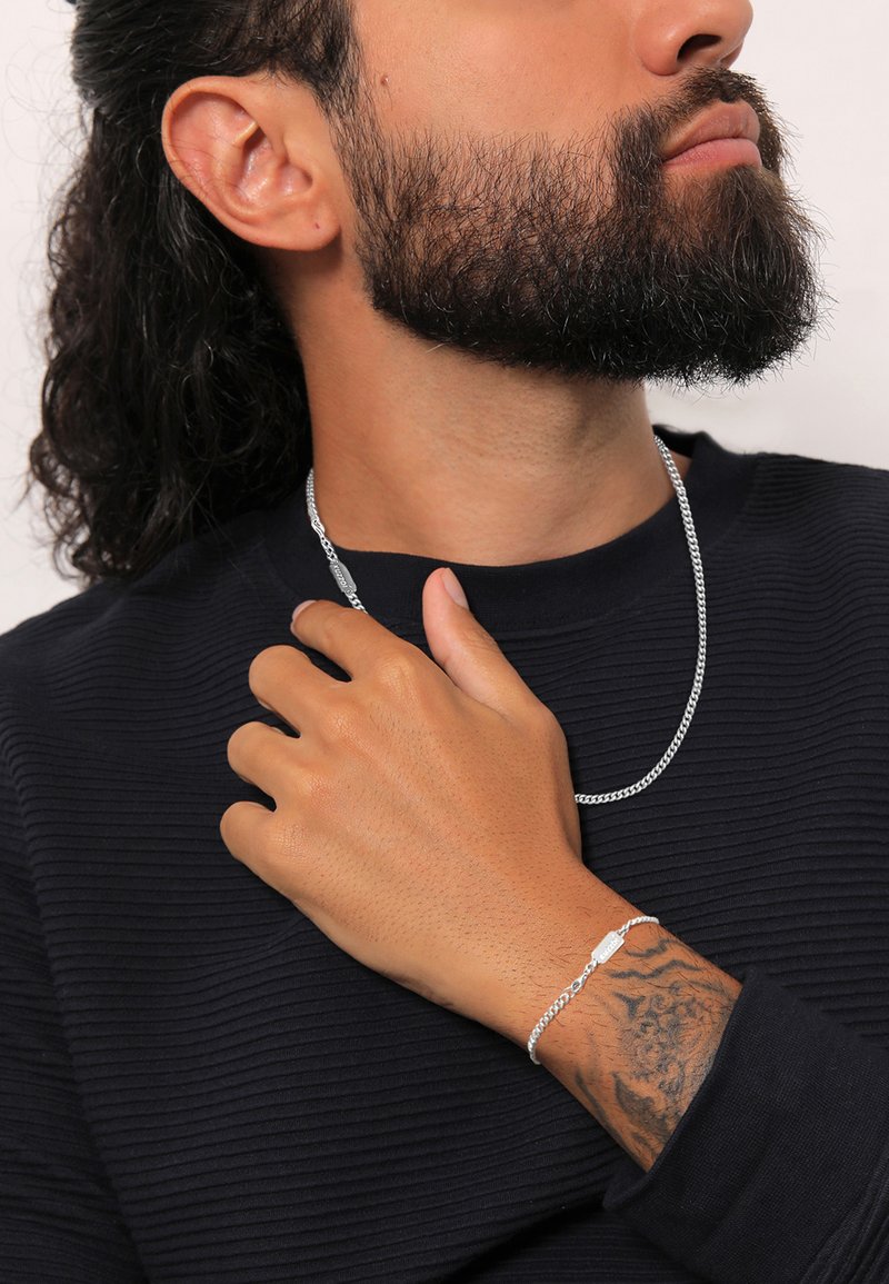 Collana e bracciale in argento su un polso di tono della pelle, indossati con una camicia nera testurizzata. La collana presenta un design semplice.