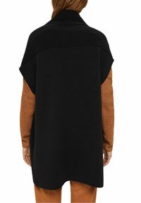 Zwarte gebreide poncho met een afgeronde zoom en oversized ontwerp, gelaagd over een textuur bruin kledingstuk met lange mouwen.