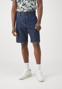 Shorts in denim di colore blu scuro. Design a gamba dritta con orlo grezzo. Abbinati a scarpe da ginnastica bianche e calze bianche a metà polpaccio.