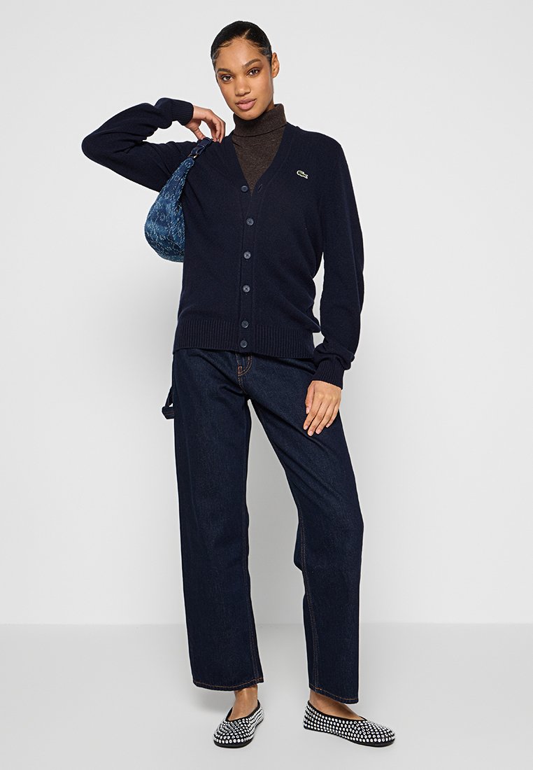 Cardigan blu navy con chiusura a bottoni e polsini a costine, indossato sopra un dolcevita marrone. Abbinato a jeans scuri e scarpe slip-on a fantasia.