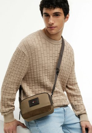Ung mand iført en beige tekstureret sweater og lyseblå jeans, siddende med en brun crossbody taske.
