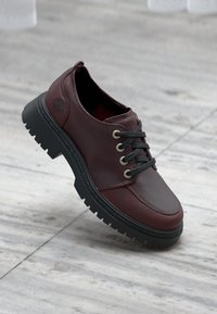 Pantofi din piele burgundy cu sireturi negre, vârful rotund și talpă groasă din cauciuc negru. Dispune de ochiuri metalice și un detaliu logo subtil pe lateral.