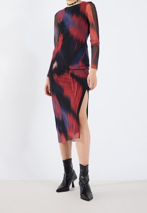 Vrouw draagt een midilange, rood, zwart en blauw tie-dye jurk met lange mouwen en een hoge split aan de dij, zwarte enkellaarsjes en een met studs versierde choker.