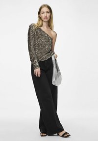 Top monospalla in bronzo metallico con paillettes abbinato a jeans neri a vita alta e a gamba larga; tiene una borsa argentata; sandali neri.