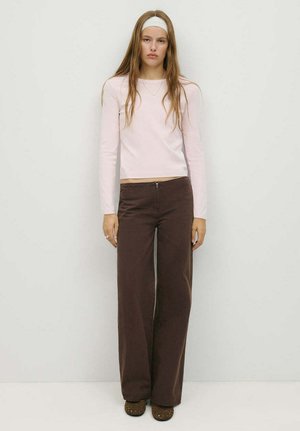 Joven con el pelo largo que lleva una cinta blanca en la cabeza, una camisa de manga larga rosa pálido, pantalones marrones de pierna ancha y zapatos planos frente a una pared lisa.