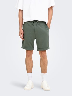 Persona con pantalones cortos verdes con cordón, camiseta blanca, calcetines blancos y zapatillas blancas, de pie con las manos en los bolsillos.