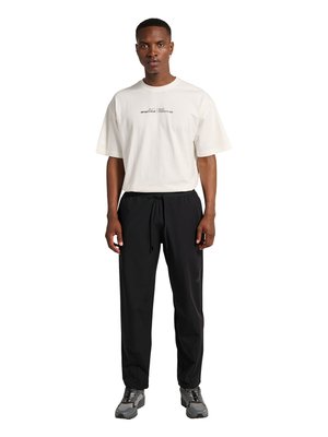 Mand iført hvid kortærmet t-shirt med tekst og sorte bukser med snøre, stående med armene afslappet ned langs siden foran en hvid baggrund.