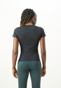 Kvinne med langt flettehår iført en tettsittende svart kortermet skjorte og mørkegrønne tights, vist fra baksiden mot en hvit bakgrunn.