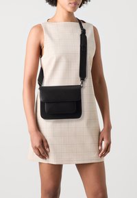 Sorte læder crossbody taske med flap-lukning, der har en glat overflade og en skulderrem, båret over en lysternet kjole.