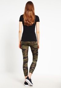 En kvinna med långt brunt hår bär en svart t-shirt och gröna kamouflage leggings, vänd bort, stående mot en enfärgad bakgrund.