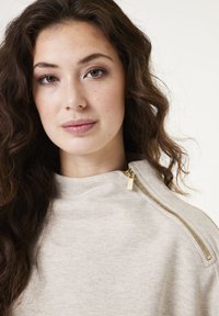 Suéter beige claro con alto cuello, que presenta un detalle de cremallera dorada en el hombro. Tejido suave y texturizado con un corte relajado.