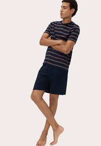Pruhované bavlněné tričko v námořnické modré, červené a bílé barvě; doplněné o námořnické šortky. Tkanina vypadá hladká; model stojí bosý se založenýma rukama.