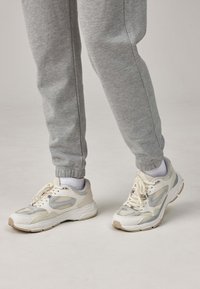 Weiße und hellgraue Sneaker mit Netzpaneelen, Schnürsenkeln und strukturierten Sohlen, kombiniert mit grauen Jogginghosen mit elastischen Bündchen.