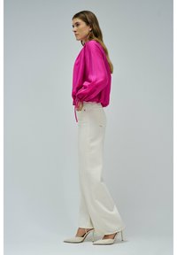 Donna con una blusa a maniche lunghe di un vivace rosa, pantaloni bianchi a vita alta e a gamba larga, e scarpe col tacco beige, in piedi di lato con le mani nelle tasche.