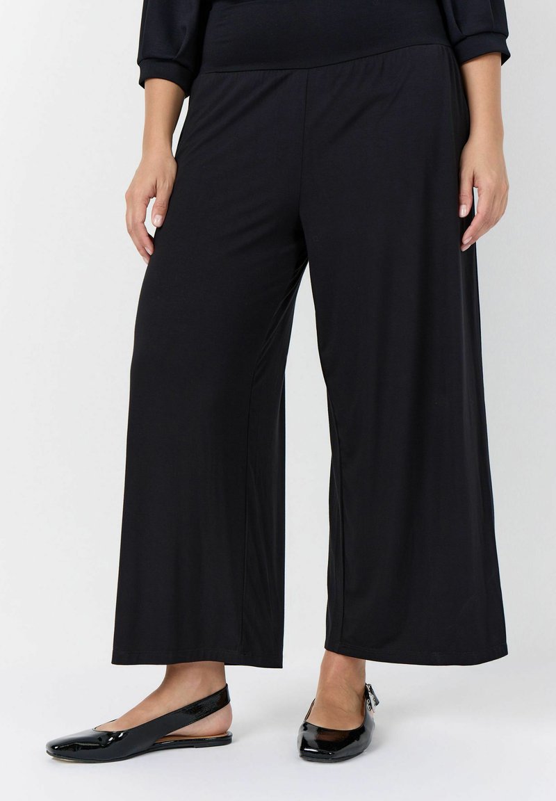 Pantalon noir à jambes larges porté avec des chaussures plates slingback en cuir verni noir, mains reposant le long du corps sur fond blanc.