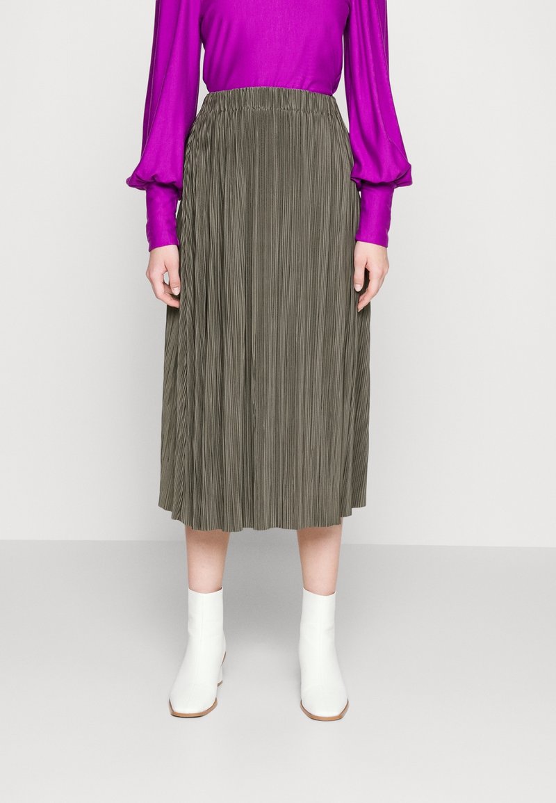 Kvinna klädd i en plisserad olivgrön midikjol, en ljuslila långärmad blus och vita ankelstövlar, stående mot en enfärgad bakgrund.