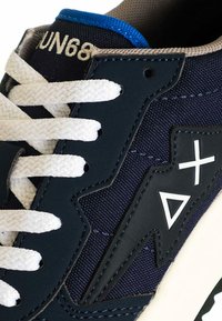 Primo piano di una sneaker blu navy con lacci bianchi, rifiniture beige e dettaglio di un fulmine nero con simboli triangolari e incroci bianchi.