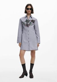 Grå skjortklänning med blommig brodyr på bröstet, knäppning framtill, långa ärmar och rak silhuett. Bärs med svarta strumpor och skor.