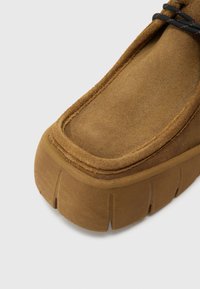 Καφέ παπούτσι από σουέτ με παχύ, υφασμένο πάτο και στρογγυλεμένη μύτη. Διαθέτει ορατές ραφές και ελαφρύ χείλος γύρω από την άκρη.