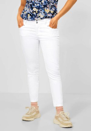 Kvinde iført hvide slim-fit bukser, blå blomstret top, lyserøde sokker og beige sneakers, stående med hænderne i lommerne.