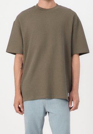 Homme portant un t-shirt ample à manches courtes côtelé de couleur vert olive, associé à un jean bleu clair, sur fond blanc uni.