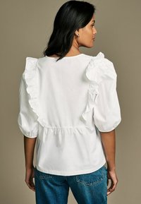 Bluză albă din bumbac, cu mâneci bufante și detalii cu volane, spate plisat și o croială lejeră, asociată cu blugi din denim albastru.