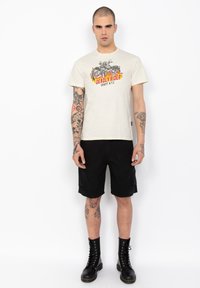 Crème korte mouwen t-shirt met een multicolor graphic en de tekst "DEADLY RIDER". Model draagt zwarte cargoshorts en zwarte veterschoenen.
