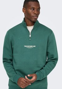 Uomo che indossa un felpa verde con zip a quarti con la scritta "THE CULTURE CAFE", in piedi con le mani incrociate e con lo sguardo di lato.