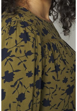 Olivově zelená halenka s černým květinovým potiskem a nařasenými detaily na rameni a na hrudi, kterou nosí osoba s kudrnatými tmavými vlasy.