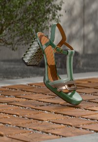 Πράσινο σανδάλι από πατενταρισμένο δέρμα με δομημένο σχήμα, μεταλλικές λεπτομέρειες και τακούνι τύπου block με σχέδιο. Διαθέτει λουράκια και κούμπωμα με αγκράφα.