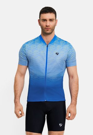 Man standing facing forward wearing a short-sleeve blue gradient cycling jersey and black cycling shorts with logos.