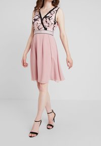 Žena v bezramínkových růžových šatech ke kolenům s černou květinovou výšivkou na vršku a černými vysokými sandály na kotníkovém pásku.