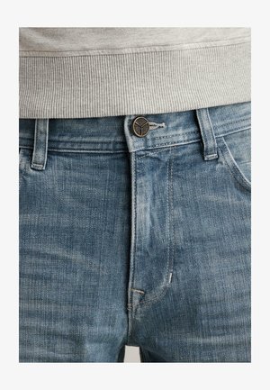 Primer plano de unos vaqueros de mezclilla azules abrochados con un botón redondo de bronce y usados con un suéter de canalé gris claro.