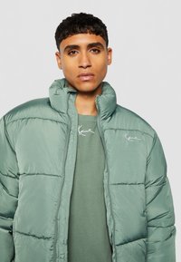 Chaqueta acolchada verde con un alto cuello, diseño acolchado y logotipo bordado en el pecho. Se lleva sobre una camisa verde a juego.