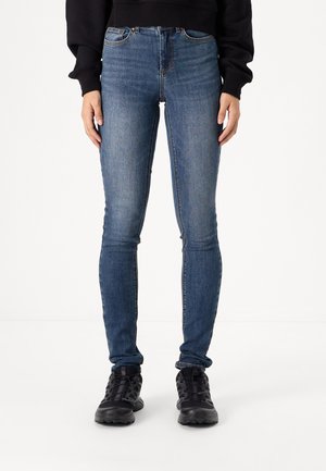 Tynne blå denimjeans med høy midje, fem-lomme design og subtile slitasjer. Brukt med svarte utendørssko.