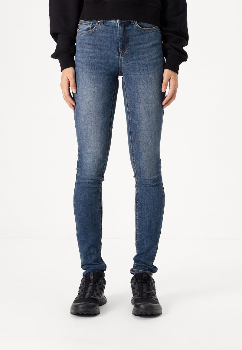 Smala blå denimjeans med hög midja, femficksdesign och subtila slitningar. Bärs med svarta utomhusskor.