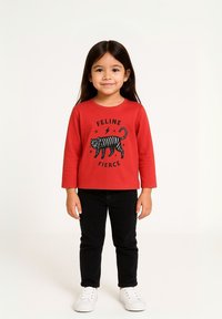 Crvena majica dugih rukava s grafikom crnog tigra i tekstom "FELINE FIERCE," u kombinaciji s crnim hlačama i bijelim tenisicama.