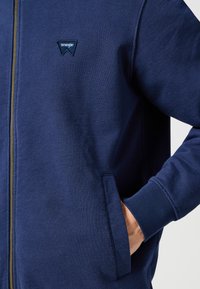 Marinblå zip-up hoodie i mjukt tyg. Har en liten broderad logga och sidofickor. Ribbstickade muddar och fåll för en tajt passform.