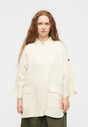 Νεαρή γυναίκα με μακριά κυματιστά μαλλιά, που φοράει χαλαρό κρεμ παλτό με κουμπιά και μεγάλες μπροστινές τσέπες, καθώς και σκούρο παντελόνι, σε απλό φόντο.