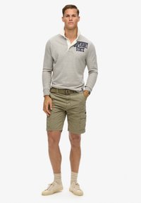 Lichtgrijze trui met een kraag, voorzien van marineblauwe tekst; groene cargoshorts; beige sneakers; zichtbaar gestructureerde stof en contrasterende lagen.