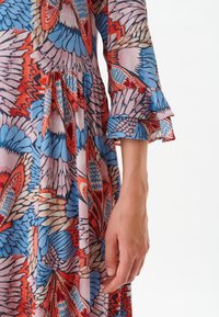 Robe colorée avec un motif floral et papillons en rose, bleu et rouge ; manches longues avec poignets à volants et texture de tissu fluide.