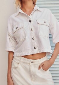 Giacca di jeans bianca a maniche corte e taglio cropped con due tasche sul petto, bottoni argentati, abbinata a una gonna bianca a vita alta.