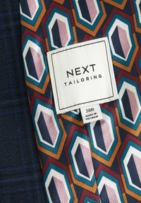 Oznaka na sako jakni s geometrijskim uzorkom u tamnoplavoj, crvenoj, narančastoj i tirkiznoj boji. Sadrži bijelu oznaku s natpisom "NEXT TAILORING" i detaljima o veličini.
