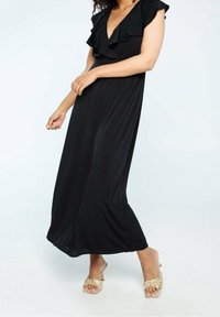 Robe maxi noire avec des manches cap ruffle, décolleté en V profond et tissu fluide. Portée avec des sandales à brides beiges, mettant en évidence une coupe décontractée.
