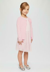 Lichtroze cardigan met knoopsluiting en bloemenaccent, laagjes witte jurk met geplooide rok, en glanzende zilveren ballerina's.